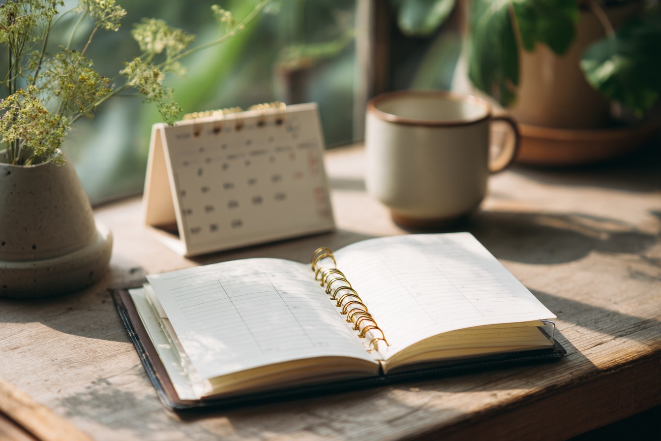 Illustrative image: an open ring planner beside a desk calendar and a mug in summer light.